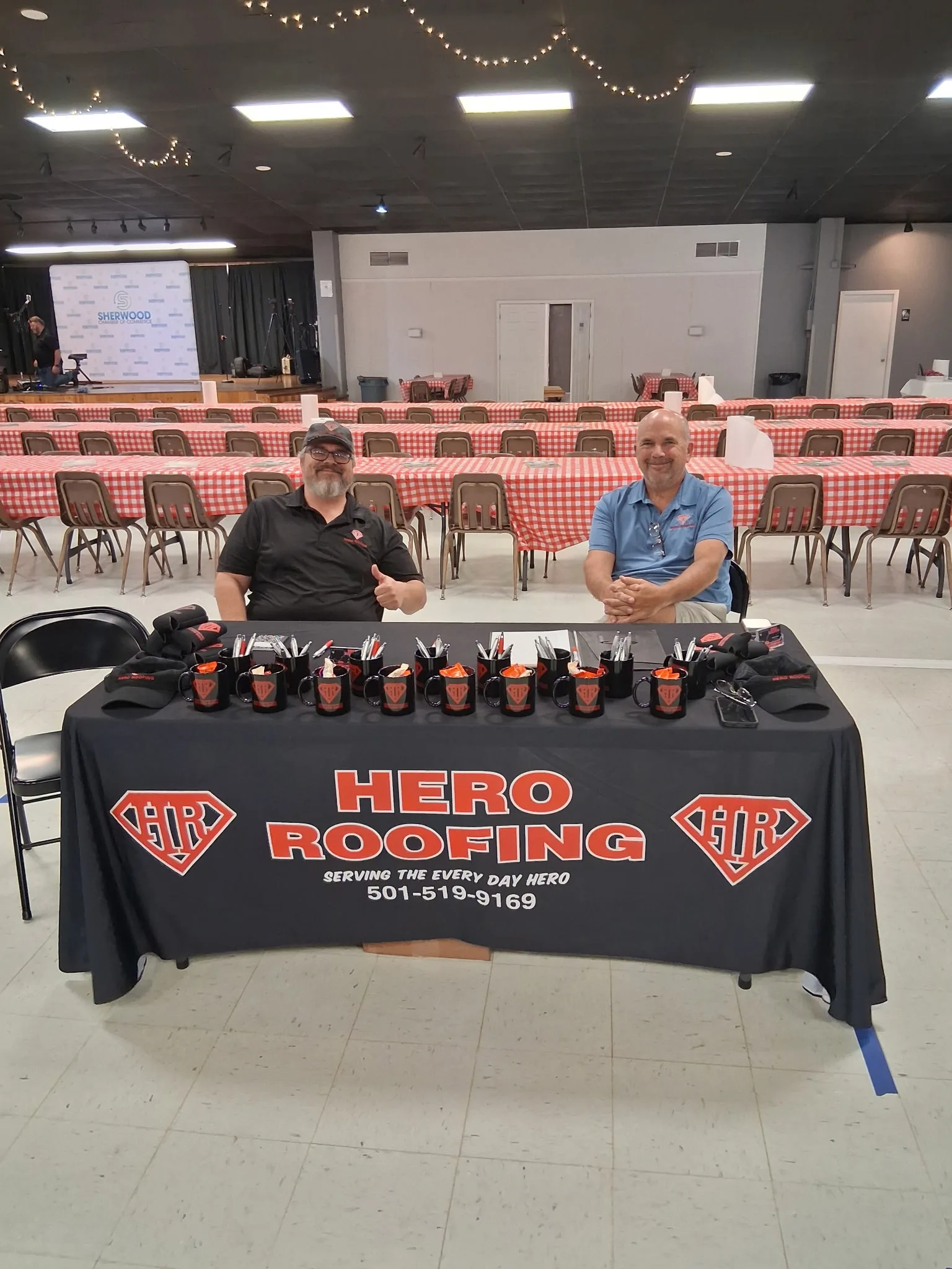 Two Hero Roofing team members seated at a table with company hats and mugs at the Sherwood Fish Fry
